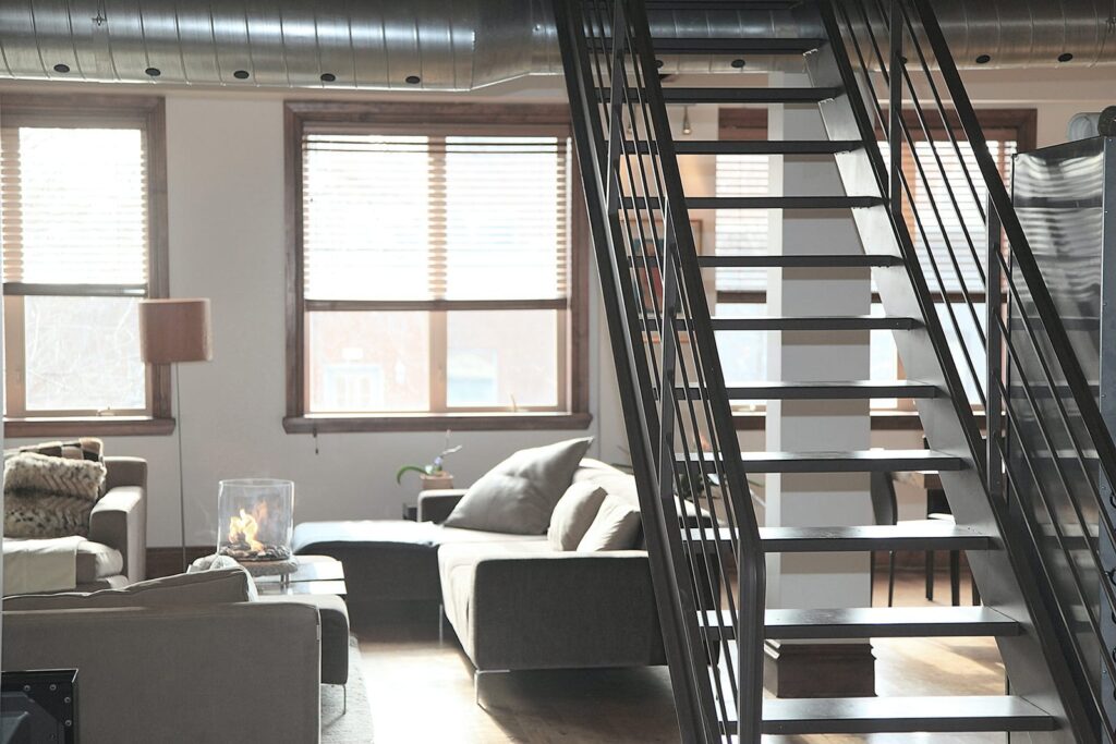 Industrial-style living room with a metal staircase on the right, gray sofas, and a glass fireplace.