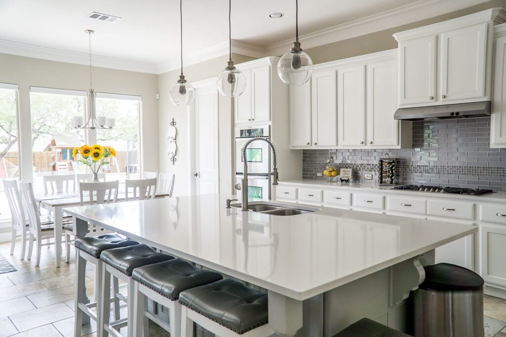 Bright white kitchen with a large island, hanging glass pendant lights, and a sink; sunflowers in a vase on the dining table by sunny windows.