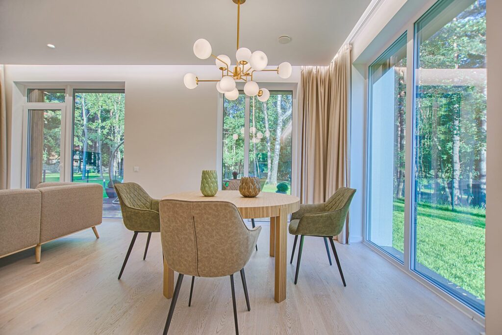 Bright dining area with a round wooden table, beige upholstered chairs, and a gold constellation chandelier, lit by large windows overlooking a green yard.